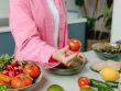 Person in pink overshirt holding a fresh tomato with various organic vegetables on a countertop.