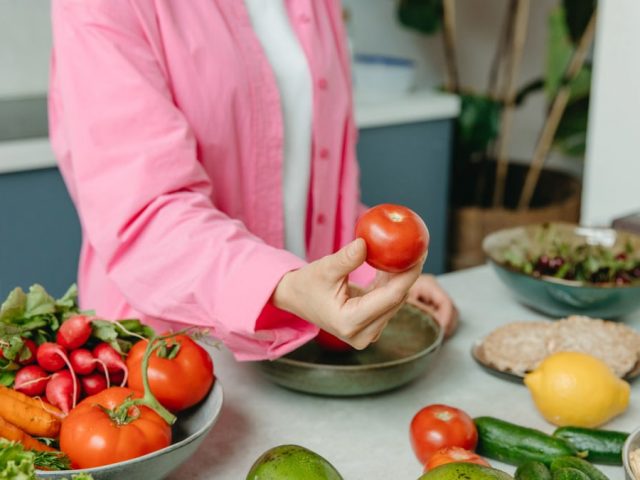 Person in pink overshirt holding a fresh tomato with various organic vegetables on a countertop.